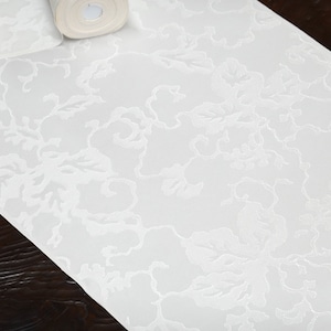May include: White table runner with an embossed floral pattern. The runner is made of a soft, textured fabric.