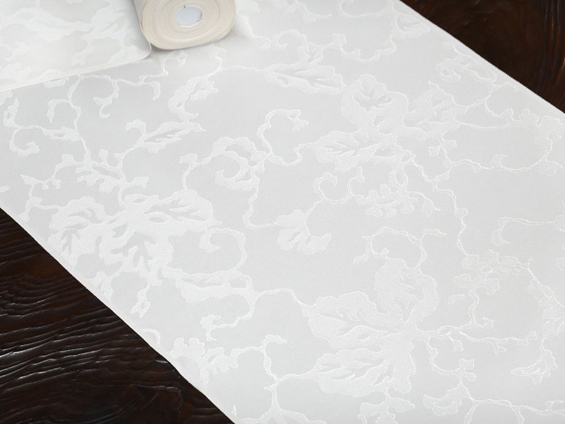 May include: White table runner with an embossed floral pattern. The runner is made of a soft, textured fabric.