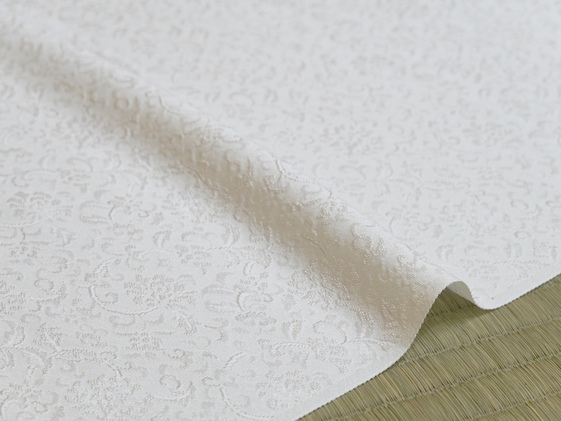 May include: White fabric with a raised, embossed pattern of small, intricate floral designs. The fabric is folded in half and has a soft, textured appearance.
