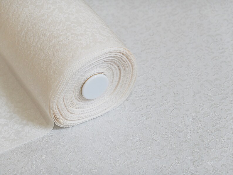 May include: A roll of white fabric with a delicate embossed pattern. The fabric is rolled up tightly and secured with a white plastic cap.