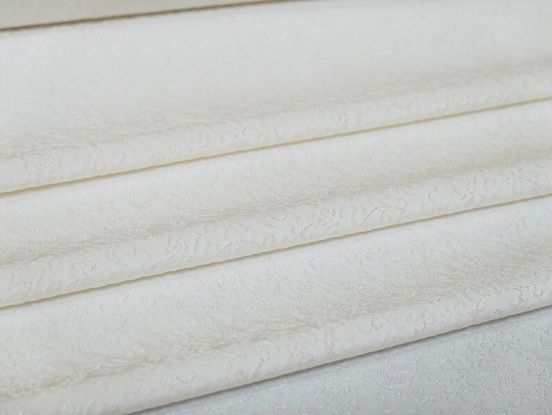 May include: White fabric with a subtle embossed pattern. The fabric is folded in layers, showcasing the texture.
