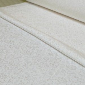 May include: White fabric with a delicate embossed floral pattern. The fabric is rolled up and folded, showing the texture of the design.