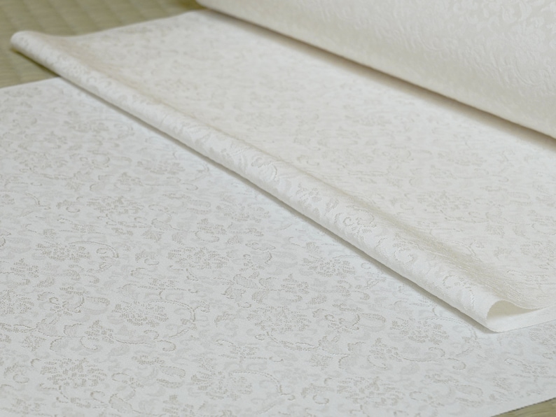 May include: White fabric with a delicate embossed floral pattern. The fabric is rolled up and folded, showing the texture of the design.