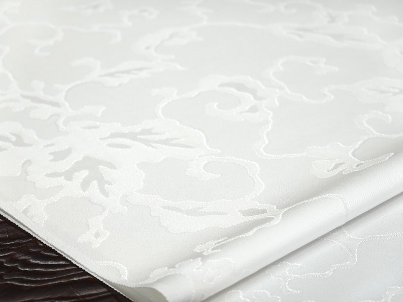 May include: White table runner with an embossed floral pattern. The runner is folded in half and has a smooth, silky texture.