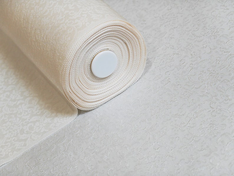 May include: A roll of white fabric with a delicate floral pattern. The fabric is rolled up and secured with a white plastic core.