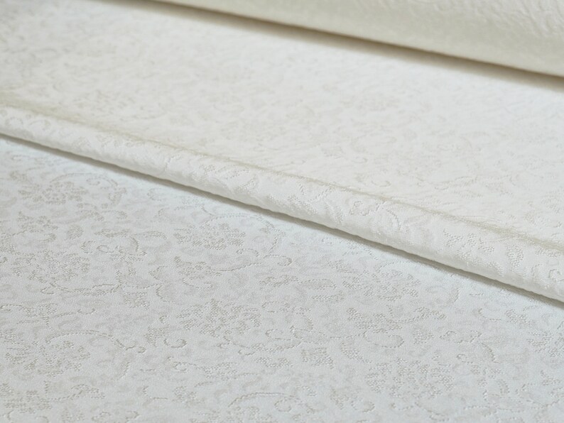 May include: White fabric with a subtle embossed floral pattern. The fabric is folded over, showing the texture of the pattern.
