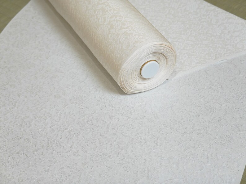 May include: A roll of white fabric with a delicate embossed pattern. The fabric is rolled up and secured with a white plastic cap.