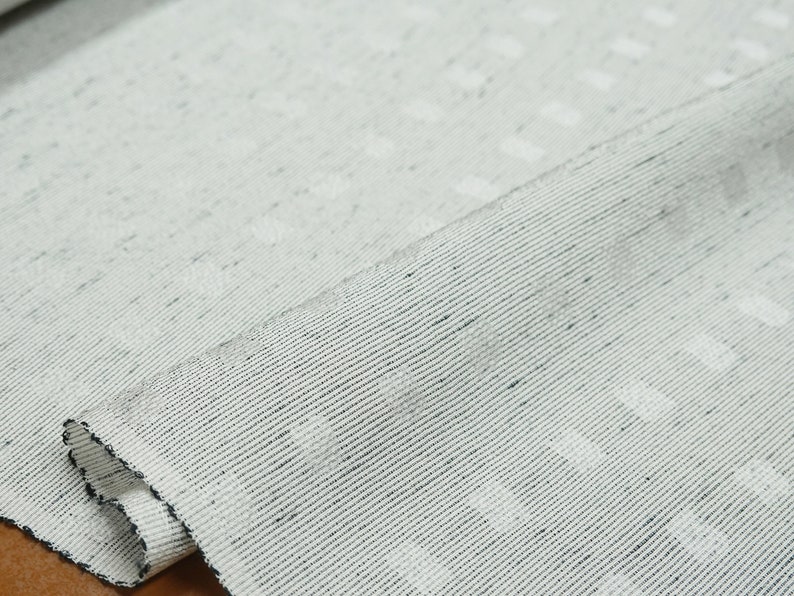 May include: Close-up of a white fabric with a subtle textured pattern. The fabric has a slightly rough, woven texture and a subtle sheen.