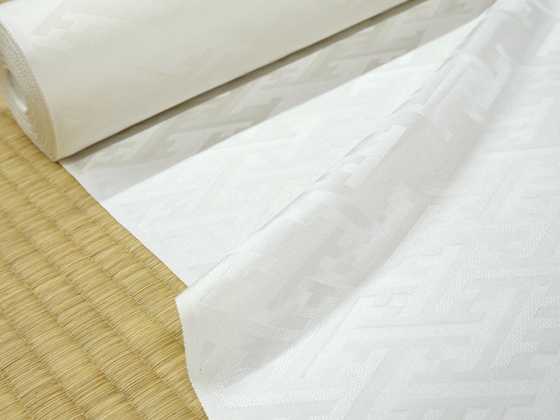 May include: White fabric with a geometric pattern, rolled up and displayed on a woven mat.