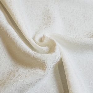 May include: White fabric with a subtle embossed floral pattern. The fabric is folded and draped, showcasing its texture and drape.