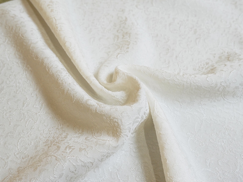 May include: White fabric with a subtle embossed floral pattern. The fabric is folded and draped, showcasing its texture and drape.