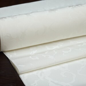 May include: A white fabric with a subtle embossed floral pattern is rolled up. The fabric is smooth and has a luxurious feel.