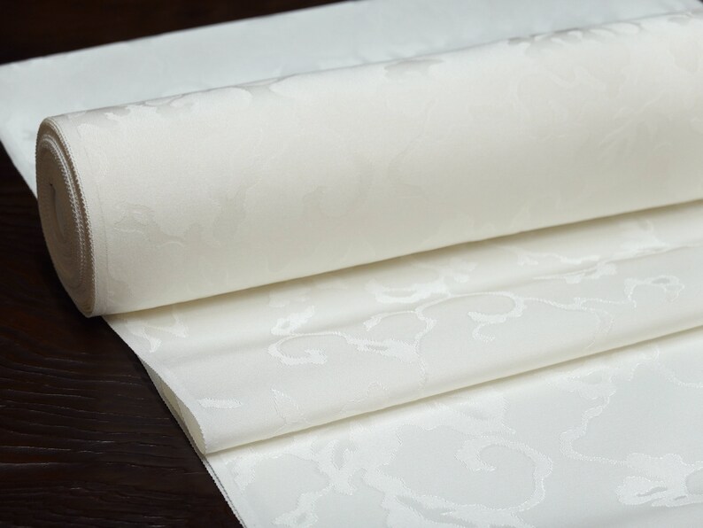May include: A white fabric with a subtle embossed floral pattern is rolled up. The fabric is smooth and has a luxurious feel.