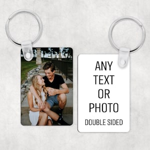 Op de afbeelding: Twee rechthoekige sleutelhangers met zilveren sleutelringen. Eén sleutelhanger toont een foto van een stel, de andere heeft de tekst "ANY TEXT OR PHOTO DOUBLE SIDED" in zwart op een witte achtergrond. De sleutelhangers zijn waarschijnlijk gepersonaliseerde cadeaus.
