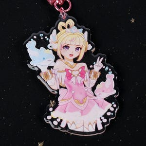 May include: A pink and white acrylic charm featuring an anime-style character with blonde hair, a pink dress, and two small dolphin figures. The charm has a pink keychain attachment and is set against a black background with glitter.