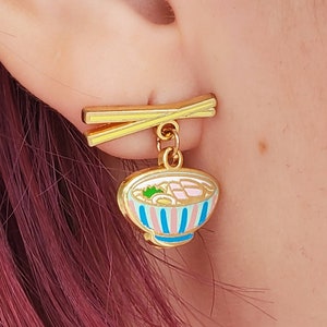 May include: Gold earrings with a ramen bowl charm. The bowl is pink, blue, and white with a yellow rim. The chopsticks are gold and have a yellow and white stripe.