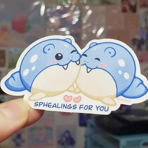 May include: A sticker featuring two cartoon seals in light blue with white glitter accents, kissing. The sticker has the text "SPHEALINGS FOR YOU" in white, with two pink hearts. The artist's signature "NIMOCHI" is in the bottom right corner.