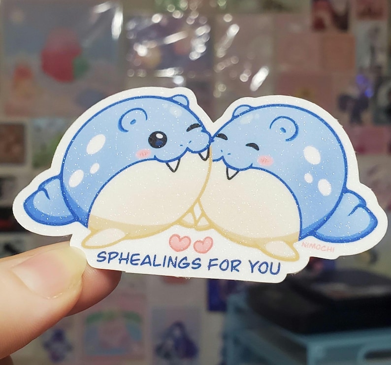 May include: A sticker featuring two cartoon seals in light blue with white glitter accents, kissing. The sticker has the text "SPHEALINGS FOR YOU" in white, with two pink hearts. The artist's signature "NIMOCHI" is in the bottom right corner.