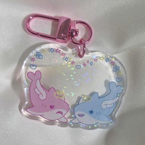 Op de afbeelding: Een doorzichtige acryl sleutelhanger met een roze haai en een blauwe haai met witte vleugels en halo's. De haaien staan tegenover elkaar, omringd door een rand van kleine kleurrijke sterretjes, hartjes en vierkantjes. Met een roze metalen sluiting.
