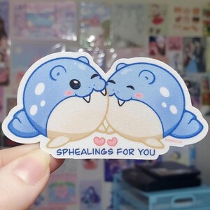 May include: A sticker featuring two cartoon seals, kissing. The seals are blue with white spots and have pink cheeks. The text "SPHEALINGS FOR YOU" is below the seals, with two pink hearts. The sticker has a white border.