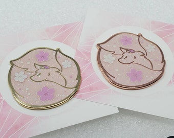 May include: Two enamel pins featuring a curled up fox with pink and white cherry blossoms. The pins are on a pink background with a geometric pattern. One pin is gold and the other is rose gold.