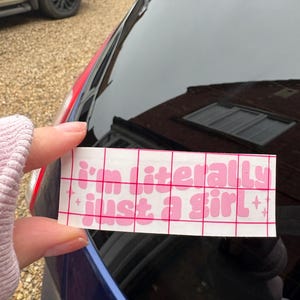Peut inclure: Un autocollant rectangulaire blanc avec le texte rose "I'm literally just a girl" en police bulle. L'autocollant est tenu contre la vitre d'une voiture.