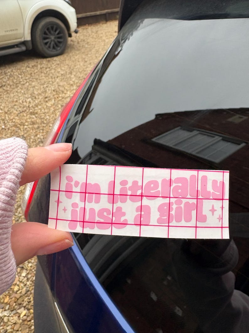 Peut inclure: Un autocollant rectangulaire blanc avec le texte rose "I'm literally just a girl" en police bulle. L'autocollant est tenu contre la vitre d'une voiture.