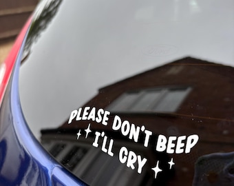 Peut inclure: Autocollant de voiture en vinyle blanc avec le texte "PLEASE DON'T BEEP I'LL CRY" dans une police ludique, orné d'étoiles. L'autocollant est appliqué sur la vitre arrière d'une voiture bleue, avec un arrière-plan flou.