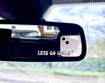 Peut inclure: Rétroviseur de voiture avec le texte "LETS GO GIRLS" dans une police rétro. Un smartphone blanc se reflète dans le miroir. L'arrière-plan montre une maison et un ciel nuageux.