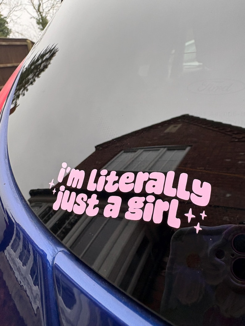 Peut inclure: Autocollant en vinyle rose avec le texte "I'm literally just a girl" dans une police r&eacute;tro, avec des &eacute;toiles. L'autocollant est appliqu&eacute; sur une vitre de voiture, refl&eacute;tant l'environnement. La voiture est bleue.