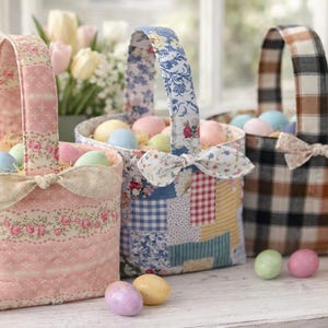 May include: Three fabric Easter baskets filled with pastel-colored eggs. The baskets have different patterns: pink floral, blue patchwork, and brown plaid. Each basket has a matching bow. A bouquet of flowers is in the background.