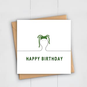 May include: A white greeting card with a minimalist illustration of a face with green hair. The words "HAPPY BIRTHDAY" are printed in green below the illustration. The card is set against a brown envelope.