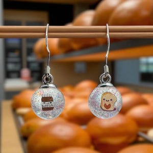 May include: Silver dangle earrings with clear, glitter-filled orbs. One orb has a Nutella jar graphic, the other a slice of toast with a heart. The earrings hang against a blurred background of baked goods.