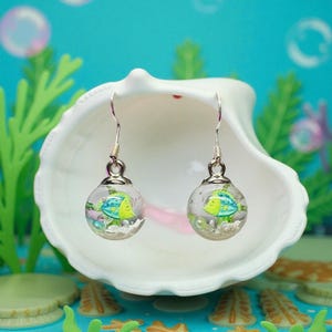 Puede incluir: Pendientes de plata con gancho. Cada pendiente presenta un globo redondo y transparente con un pequeño diseño de pez colorido en tonos azules y amarillos. Los pendientes se exhiben en una concha blanca sobre un fondo azul.