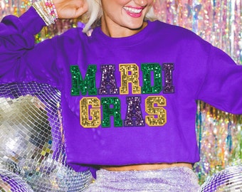 May include: Purple sweatshirt with the text "Mardi Gras" in green, gold, and purple sequins. The sweatshirt is worn by a person with a white headband and is sitting in front of a silver disco ball.