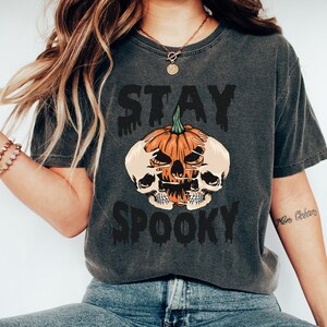 以下が含まれることがあります： ハロウィーンをテーマにしたグラフィックのダークグレーのTシャツ。デザインは、スカルと「STAY SPOOKY」の文字が滴るフォントで描かれたカボチャが特徴です。このシャツは、快適でカジュアルなスタイルです。