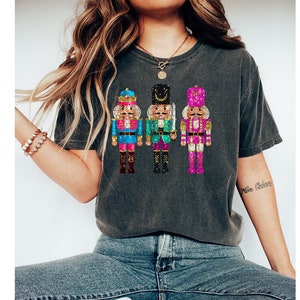May include: A dark grey t-shirt featuring a design of three nutcracker soldiers in different colours. The nutcrackers are wearing blue, green, and pink outfits with gold accents.