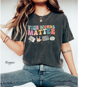 May include: A dark gray t-shirt with the text "YOUR WORDS MATTER" in rainbow colors. The shirt features a variety of colorful icons, including a heart, a hand gesture, and a phone.
