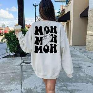 Pode incluir: Uma camisa de moletom branca com uma estampa gr&aacute;fica preta da palavra "MOH" repetida tr&ecirc;s vezes. O gr&aacute;fico tamb&eacute;m inclui uma m&atilde;o com o dedo do meio estendido.