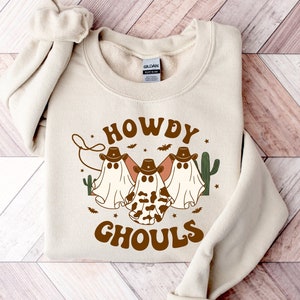 May include: Beige crewneck sweatshirt with a graphic that says "HOWDY GHOULS". The design features three ghost figures wearing cowboy hats, a cactus, and bats. The sweatshirt is made of a soft material.