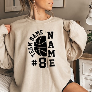 Puede incluir: Una sudadera beige con un gráfico de baloncesto negro y el texto "TEAM NAME #8 NAME" impreso en la parte delantera.
