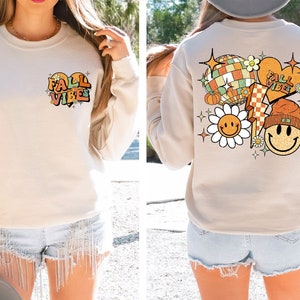 May include: A beige sweatshirt with a graphic print of a heart with the text "Fall Vibes" on the front and a graphic print of a smiley face, a heart with the text "Fall Vibes", a lightning bolt, a pumpkin, a disco ball, and a flower on the back.