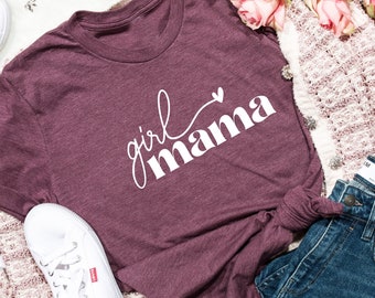 May include: A maroon t-shirt with the text "girl mama" in white lettering with a small heart above the word "mama". The shirt is laid out on a white surface with a pair of white trainers, a pair of blue jeans, and a white and pink cardigan.