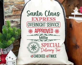 Puede incluir: Un saco de Santa Claus de lona blanca con cordón de ajuste negro. El saco tiene texto rojo y verde que dice "Santa Claus Express Overnight Service Approved Millie Special Delivery For Checked Twice".