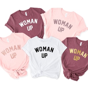 May include: Five t-shirts with the text "WOMAN UP" printed on them. The shirts are in different colors: maroon, pink, white, and two shades of pink. The text is in different colors: white, gold, and black.