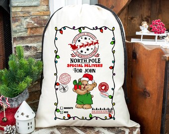 Puede incluir: Un saco de Santa de lona blanca con un cordón de ajuste negro. El saco tiene un gráfico rojo y negro con el texto "North Pole Special Delivery for John" y un oso de dibujos animados con un gorro de Papá Noel. El gráfico está rodeado de una cadena de luces navideñas.