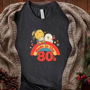 Puede incluir: Una camiseta gris oscuro con un arco&iacute;ris y el texto "Made in the 80's". El arco&iacute;ris tiene una ni&ntilde;a de dibujos animados y una criatura blanca y esponjosa encima.