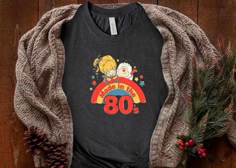 Puede incluir: Una camiseta gris oscuro con un arco&iacute;ris y el texto "Made in the 80's". El arco&iacute;ris tiene una ni&ntilde;a de dibujos animados y una criatura blanca y esponjosa encima.