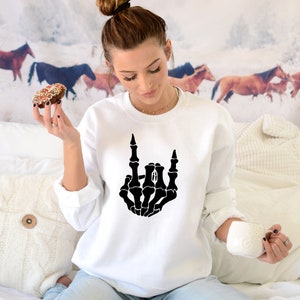 Puede incluir: Sudadera blanca con una mano de esqueleto negra haciendo el gesto de rock and roll. El gr&aacute;fico est&aacute; centrado en la parte delantera. La persona de la imagen sostiene un donut y una taza blanca.