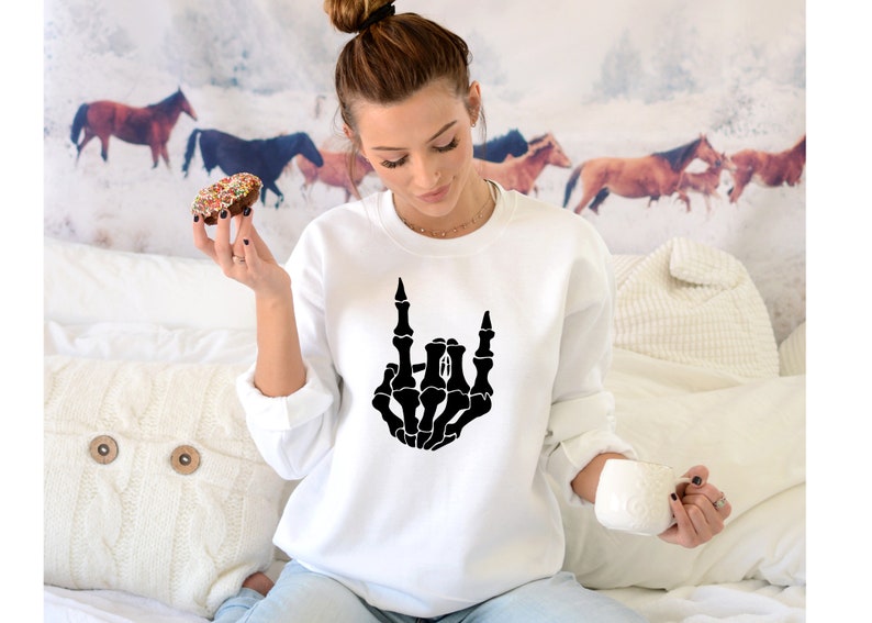 Puede incluir: Sudadera blanca con una mano de esqueleto negra haciendo el gesto de rock and roll. El gr&aacute;fico est&aacute; centrado en la parte delantera. La persona de la imagen sostiene un donut y una taza blanca.